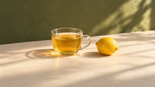 Glaskop med gylden citron te og en hel citron på et lyst bord i sollys