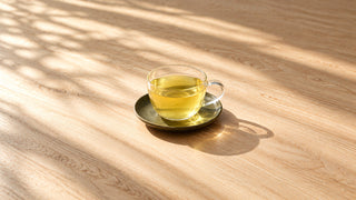 Glaskop med lysegrøn sencha på en grøn underkop på et træbord i sollys